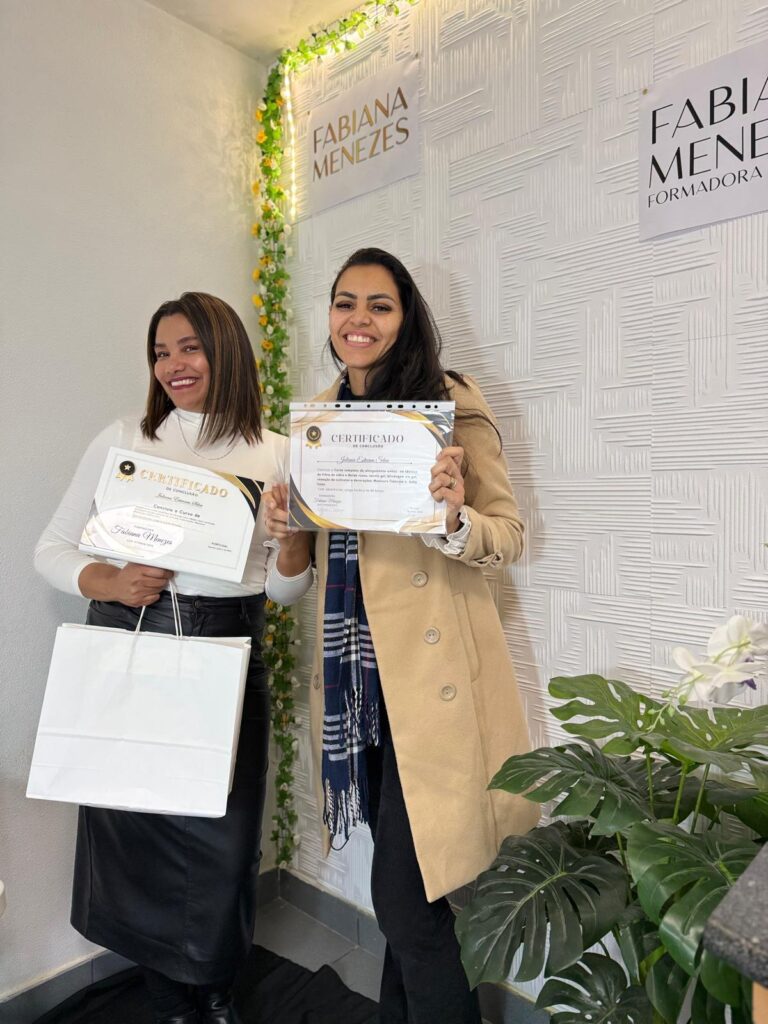 Duas alunas com os certificados, junto à parede com o nome Fabiana Menezes